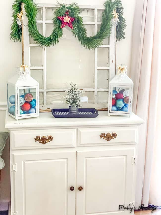 white hutch with lanterns filled with red, blue and white ornaments and greenery hung from an old window