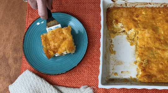 Slice of corn pudding on blue plate with white casserole dish