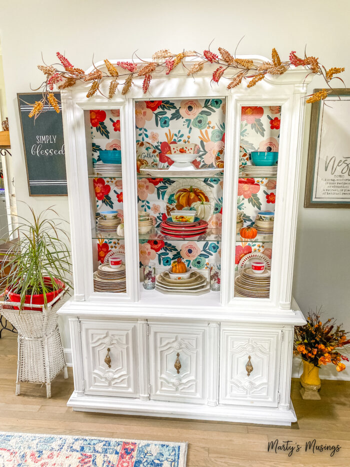 White china hutch with floral wallpaper and fall decor inside